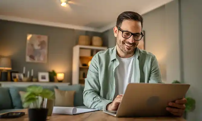 A man is sitting at a wooden table in a cozy, modern living room, smiling while using a laptop. He wears glasses and a light green jacket over a white shirt, with a few houseplants visible on the table.
