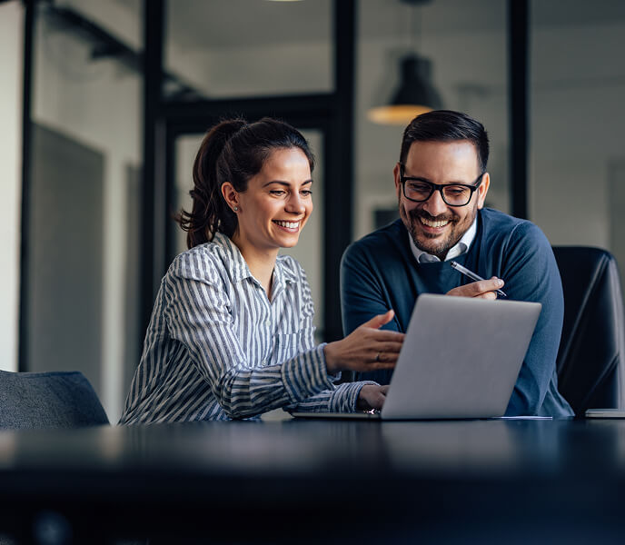 Two colleagues smiling and discussing work on a laptop in a modern office setting.