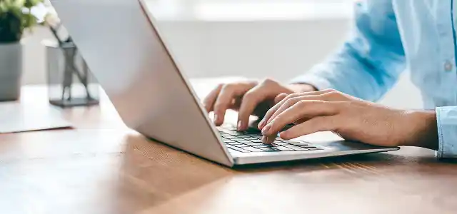 A person typing on a laptop at a wooden desk.